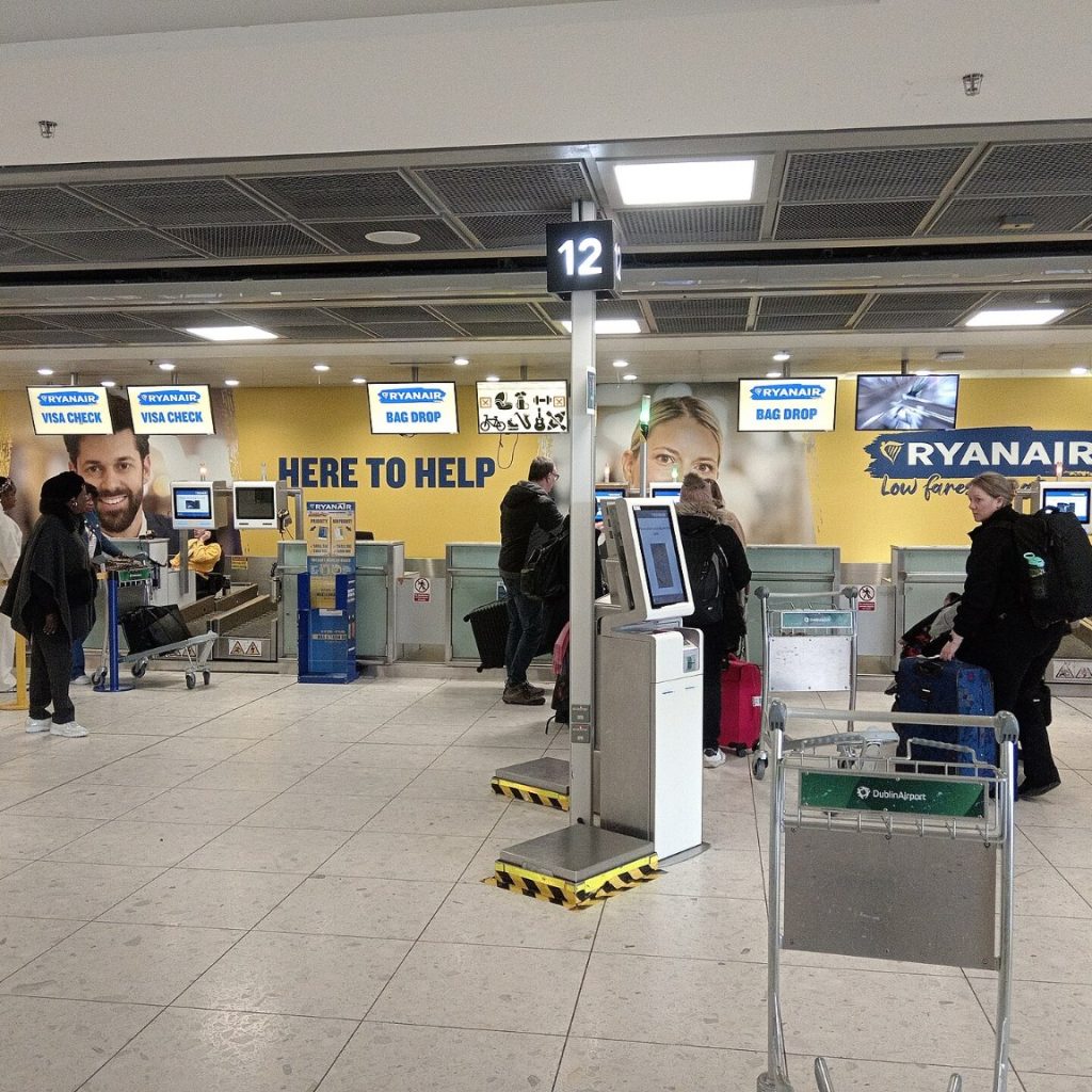 Odbavovací přepážky Ryanairu v hale letiště, před nimi samoobslužné kiosky a cestující se zavazadly