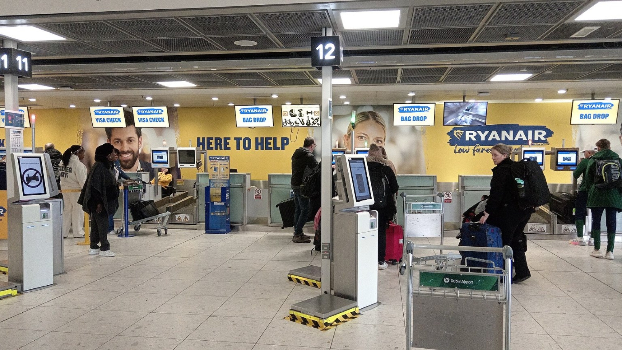Odbavovací přepážky Ryanairu v hale letiště, před nimi samoobslužné kiosky a cestující se zavazadly