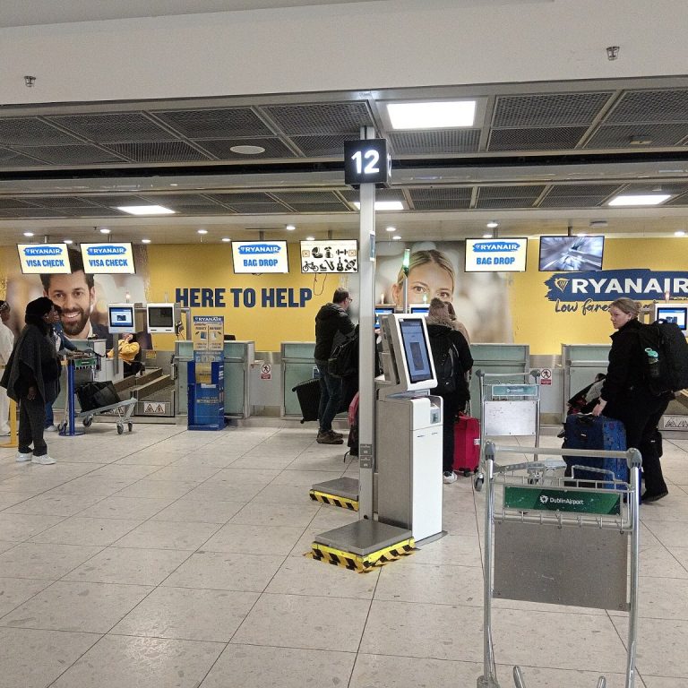 Odbavovací přepážky Ryanairu v hale letiště, před nimi samoobslužné kiosky a cestující se zavazadly