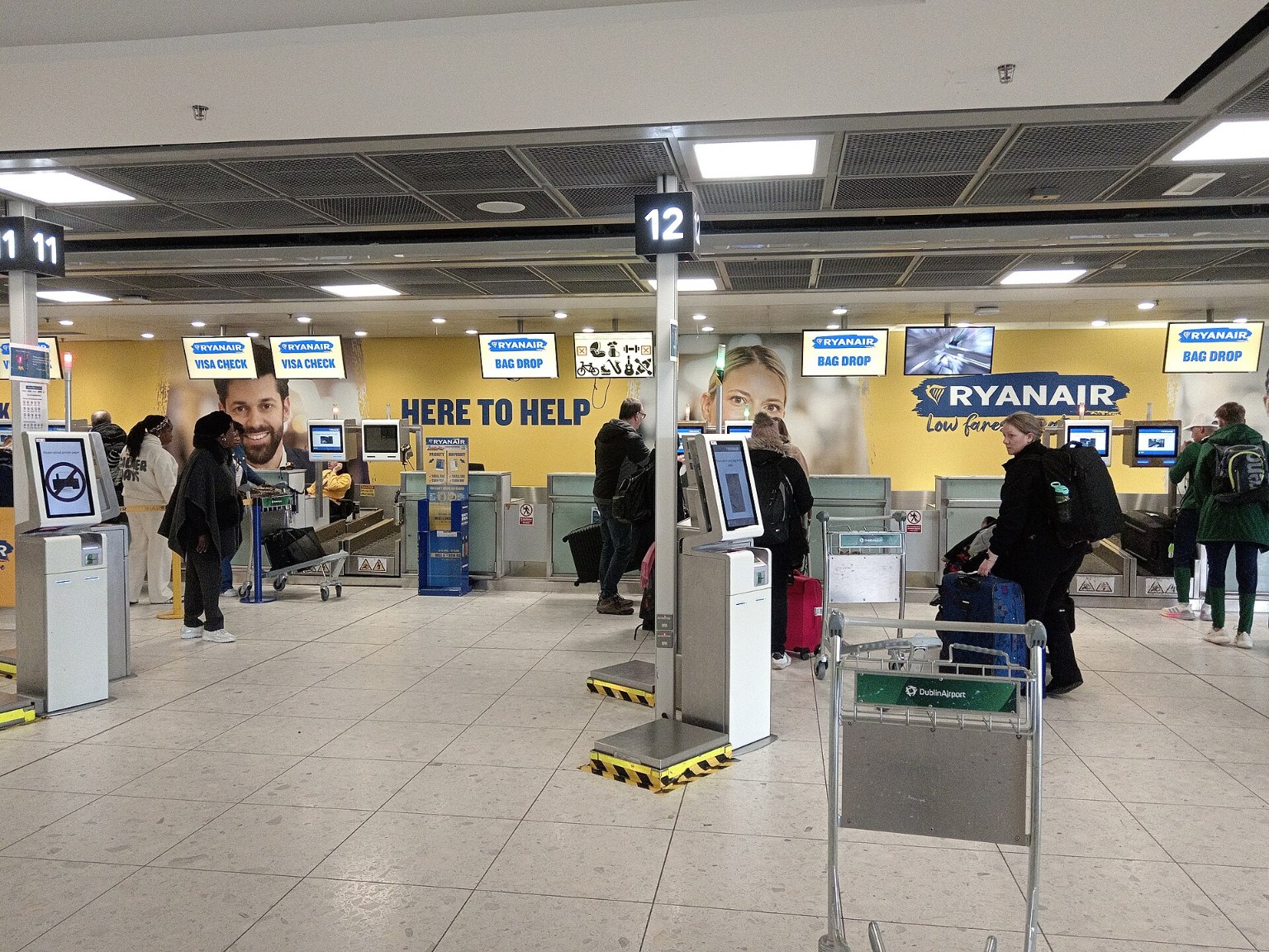 Odbavovací přepážky Ryanairu v hale letiště, před nimi samoobslužné kiosky a cestující se zavazadly